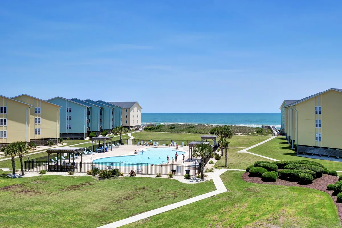 A bright outdoor scene featuring a sparkling swimming pool surrounded by lounge chairs, lush landscaping, and glimpses of the beach and ocean in the background. The pool area looks inviting for both relaxation and socializing, with a sense of privacy provided by the resort layout.