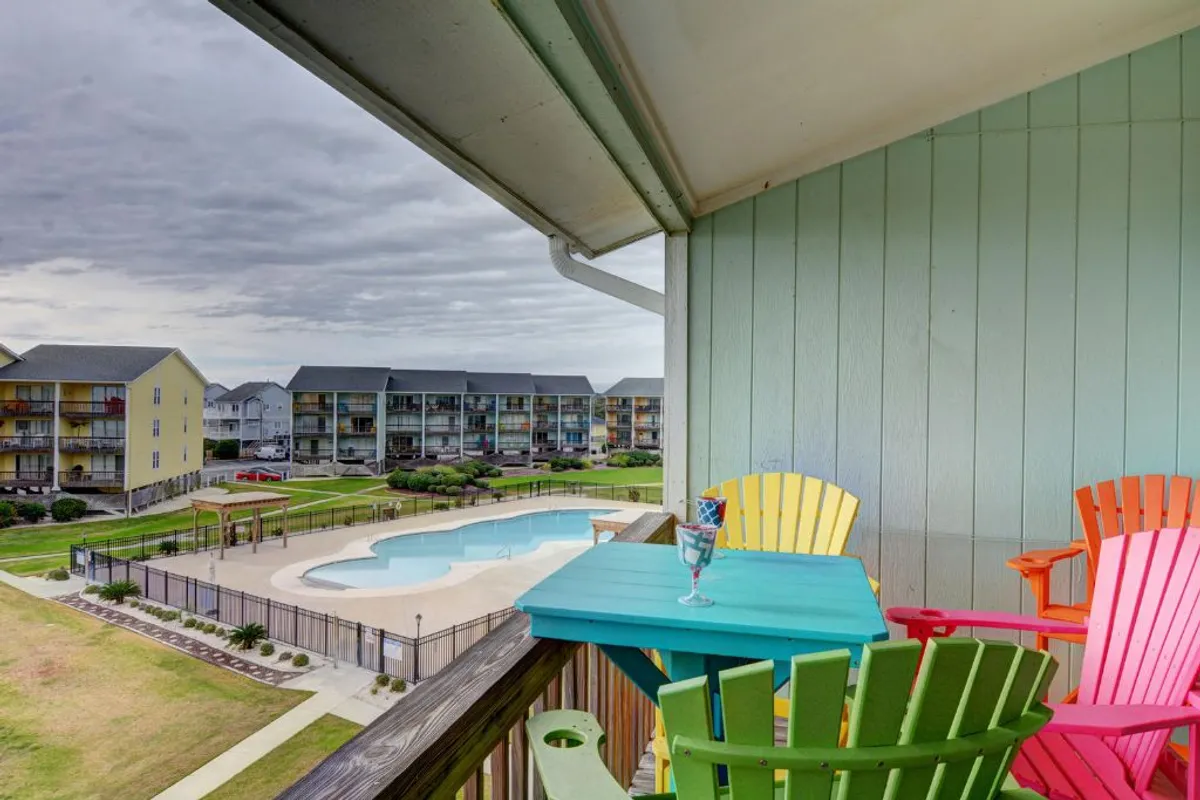 An exterior perspective showing colorful chairs set up along a pool area with a balcony railing in the foreground. The scene feels cheerful and welcoming, inviting guests to unwind, enjoy sunset views, or gather with others in the communal space.