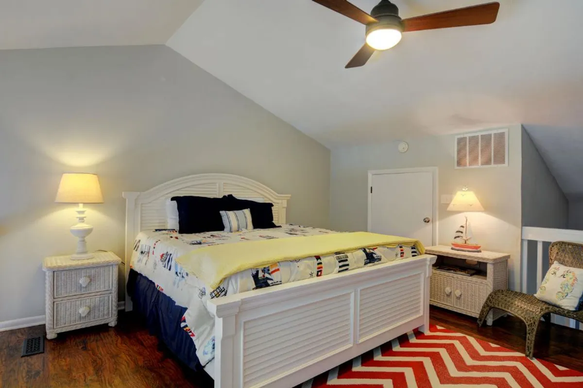 A peaceful bedroom with a colorful bedspread, a nightstand with a lamp, a striped rug, and a ceiling fan. Soft morning light fills the room, creating a calm and welcoming retreat perfect for relaxation after a day at the beach.