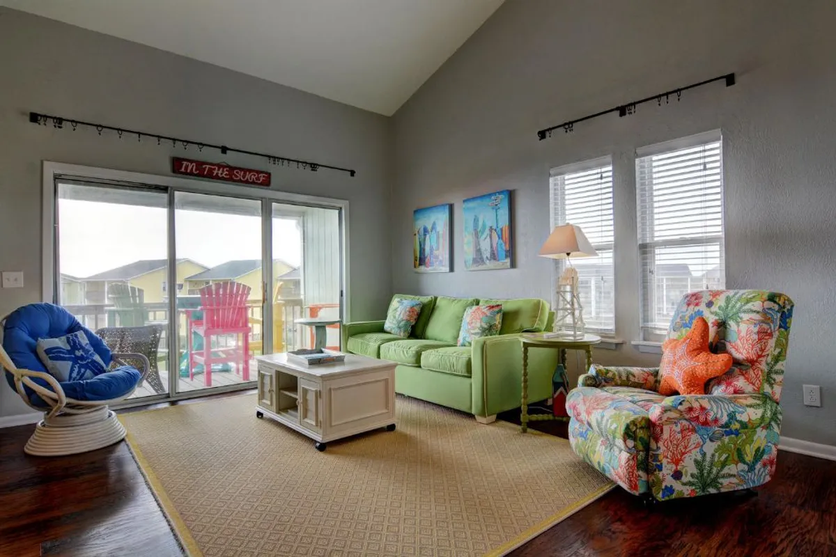 This image features a green couch and a colorful floral armchair in a bright, open living area. A large glass sliding door leads directly to a balcony, letting in ample natural light and offering a clear view of the ocean beyond. The room is adorned with brightly colored artwork, creating a playful, inviting coastal vibe.