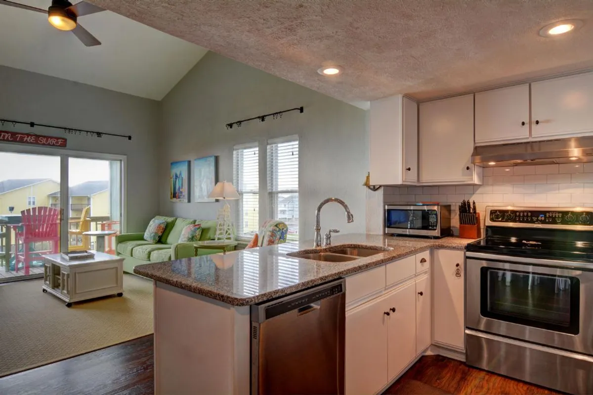 A welcoming, cheerful open living space with a kitchen island, granite countertops, stainless steel appliances, and a colorful couch. Large windows fill the room with sunlight and offer glimpses of the outdoors, reinforcing the sense of space and connection to the ocean.
