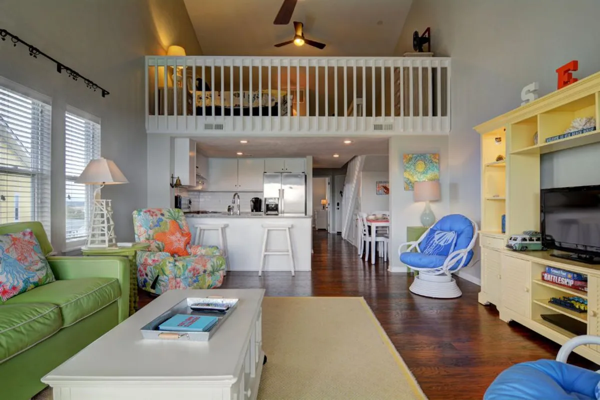 An open lofted sleeping area with a king-size bed, bright furnishings, and large windows that flood the space with sunlight. The view includes a kitchen island and family living space below, emphasizing the flexibility and spaciousness unique to this condo's layout.