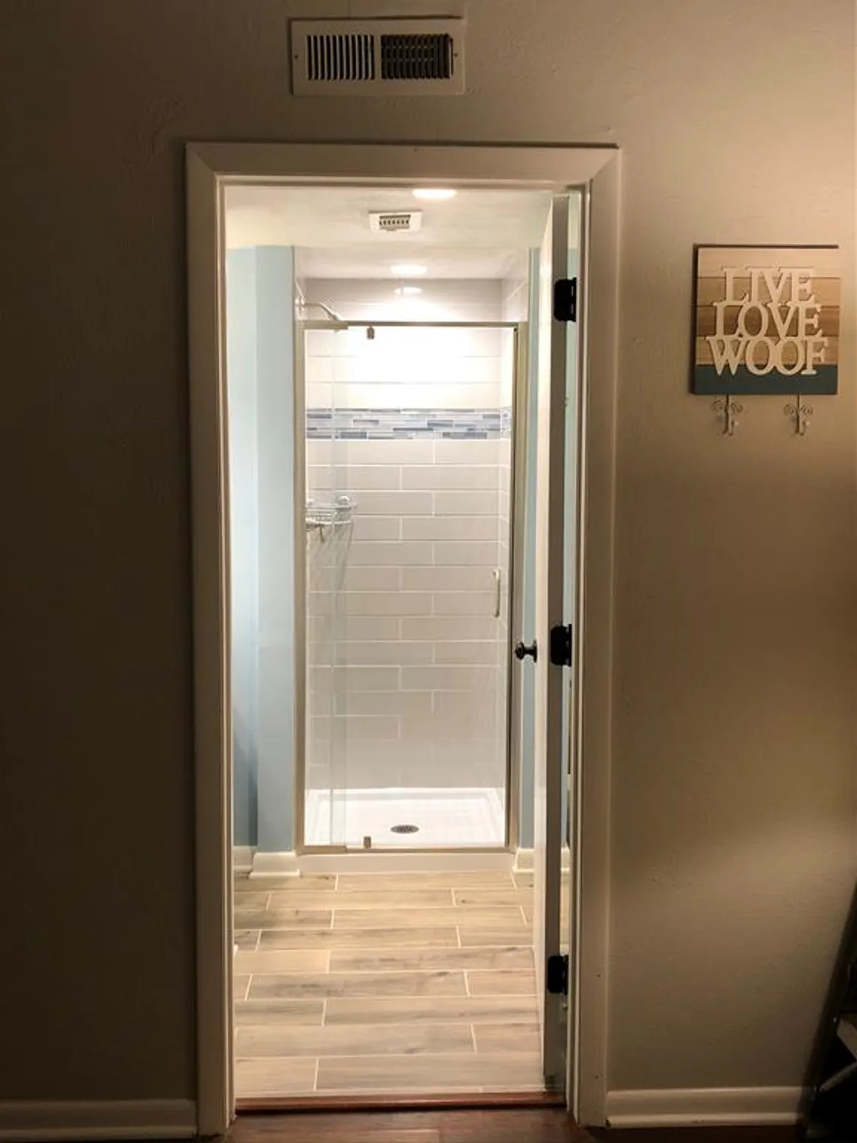 A modern bathroom featuring a stylish shower area, wood-like tile flooring, and a decorative wall sign. The space is clean and inviting, offering a rejuvenating retreat for self-care routines and relaxation.