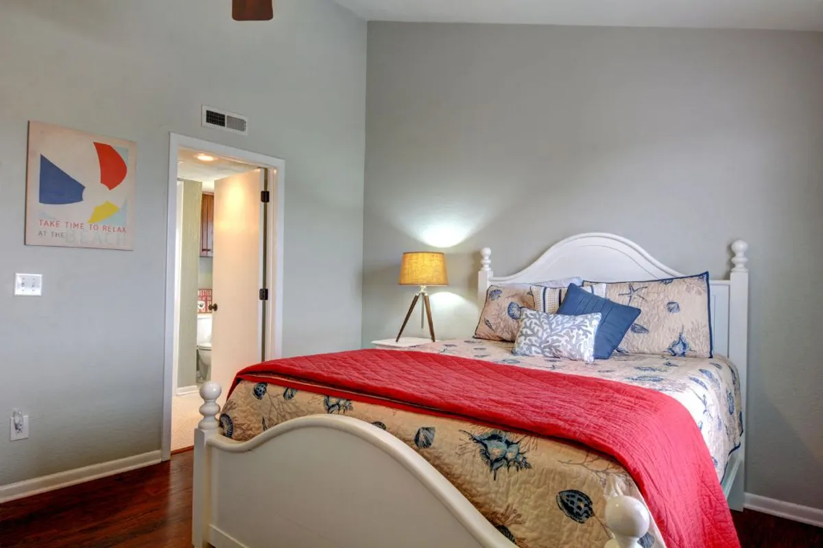 A cozy bedroom featuring a neatly made bed with decorative pillows, a bedside table with a lamp, and a piece of calming artwork on the wall. Natural light spills across the bedding, emphasizing the room’s serene atmosphere and inviting comfort.